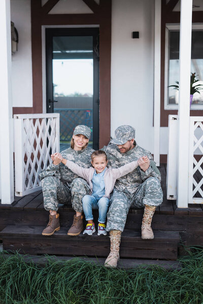 Happy daughter with outstretched hands sitting near mother and father in military uniforms on wooden threshold