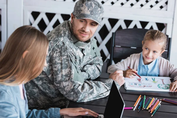 Smiling man in military uniform looking at woman typing on laptop, while sitting with daughter drawing at table