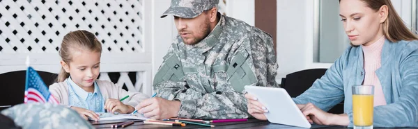 Military serviceman sitting near girl drawing and woman using digital tablet at home, banner