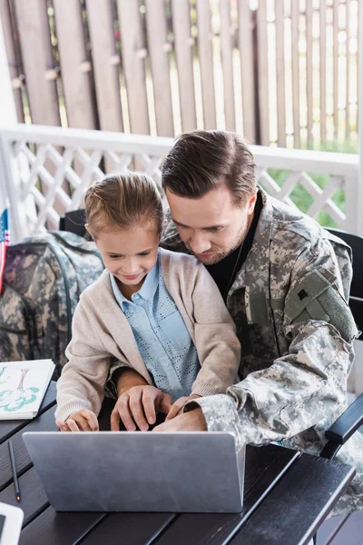 Military serviceman hugging girl and looking at laptop, while sitting at table