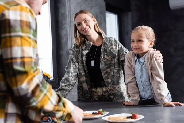 Woman in military uniform hugging daughter, while leaning on table with pancakes plates with blurred man on foreground
