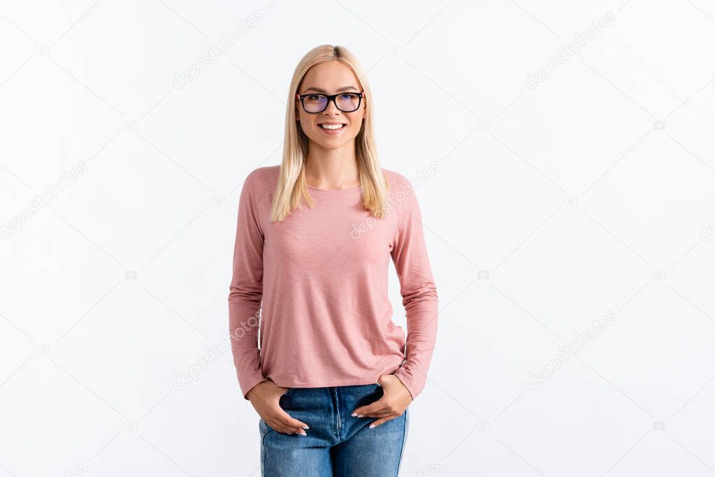 Front view of happy blonde woman with hands in pockets, wearing eyeglasses and looking at camera isolated on white