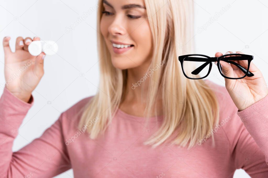 Eyeglasses in hand of smiling woman holding container with contact lenses on blurred background isolated on grey
