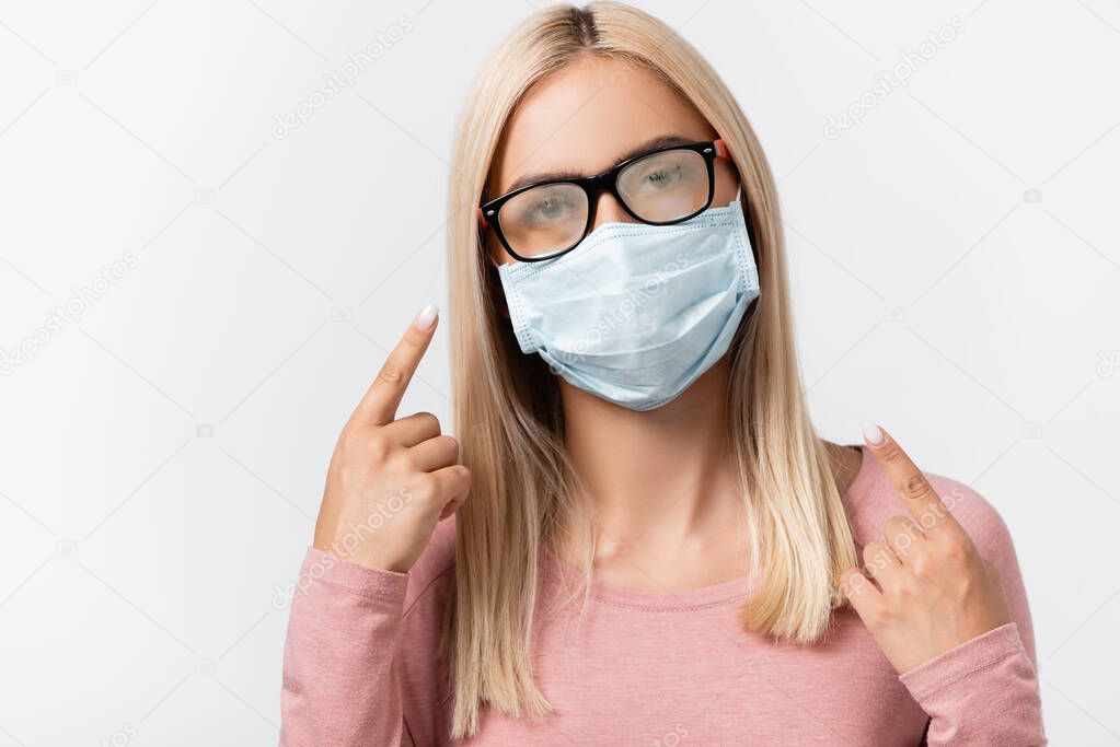 Young woman in medical mask pointing with finger at misted eyeglasses isolated on grey