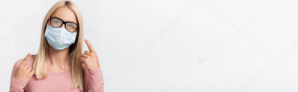 Blonde woman in medical mask pointing at misted eyeglasses isolated on grey, banner 