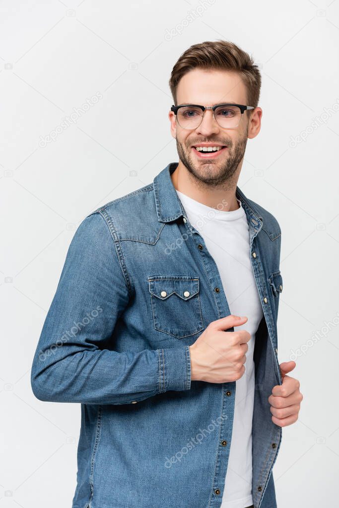 Young man in eyeglasses smiling while touching denim shirt isolated on grey