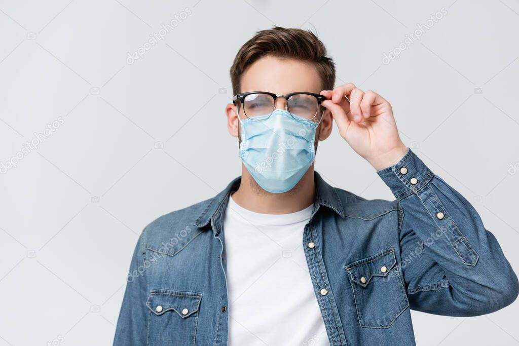 Young man in medical mask touching misted eyeglasses isolated on grey