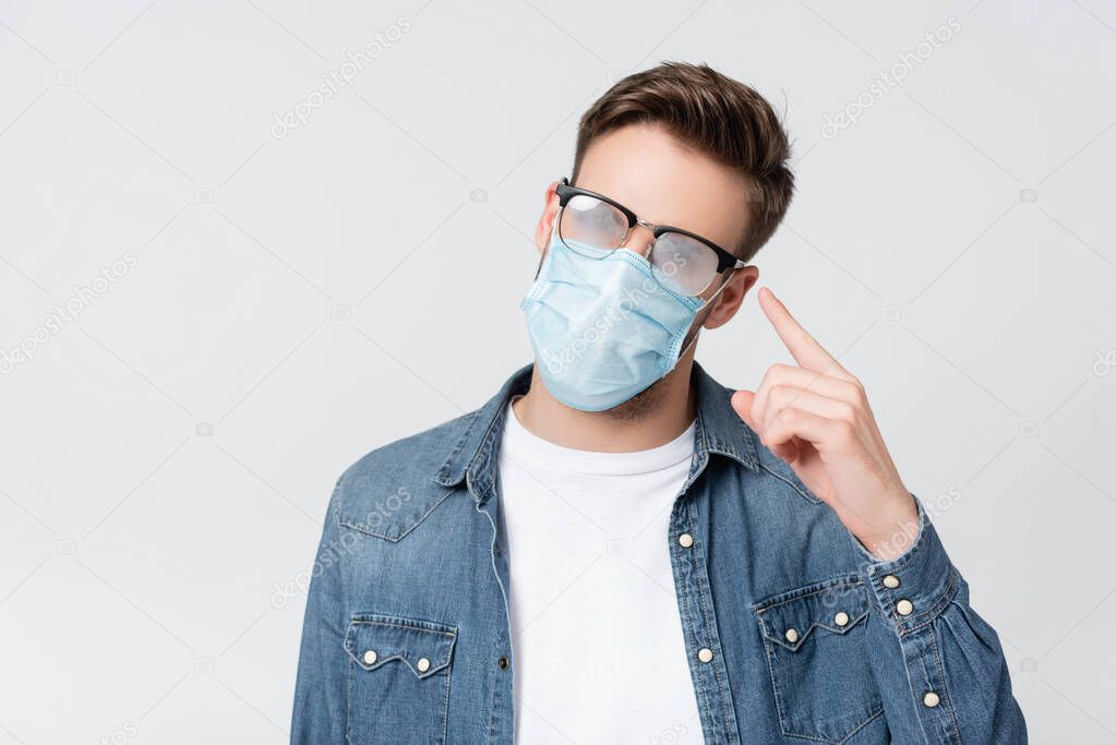 Young man in medical mask pointing with finger at misted eyeglasses isolated on grey