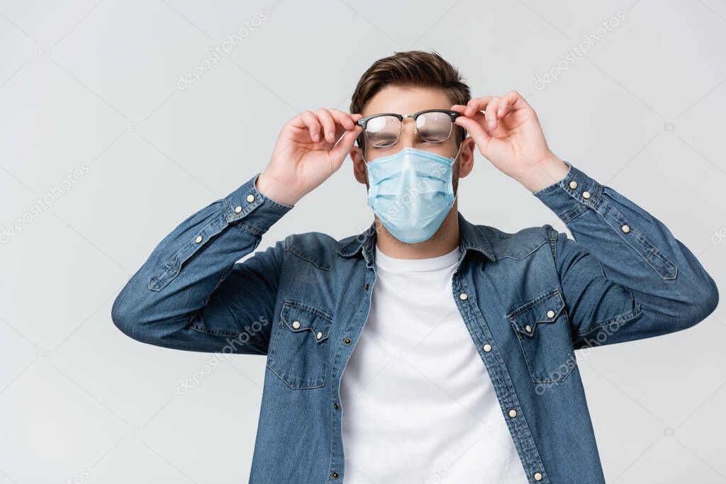 Young man in medical mask holding misted eyeglasses isolated on grey