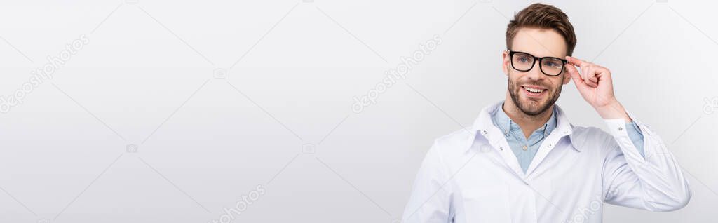 Smiling doctor touching eyeglasses while looking away isolated on grey, banner 