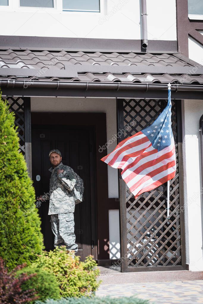 Serious military man standing near house door and looking away near bushes and american flag