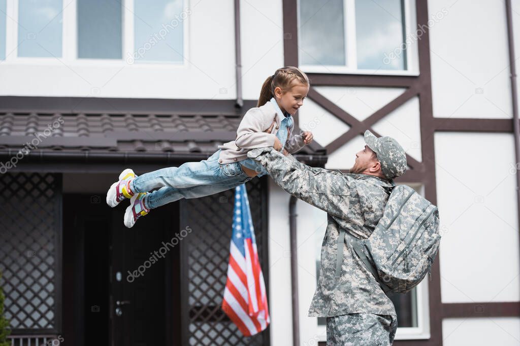 Military man lifting smiling daughter in air near house with american flag