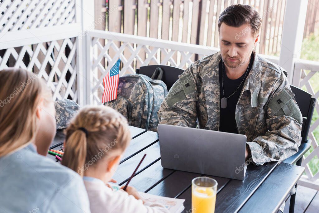Military serviceman with necklace using laptop, while sitting at table with blurred woman and daughter on foreground