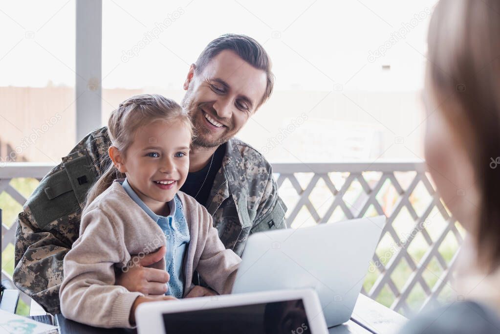 Happy girl sitting with military serviceman near laptop with blurred woman on foreground