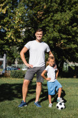 Usmívající se otec s rukama v bok s pohledem na kameru s úsměvem syn stojí s nohou na míči v parku