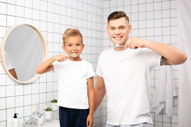 Cheerful father and son with toothbrushes looking at camera in bathroom clipart