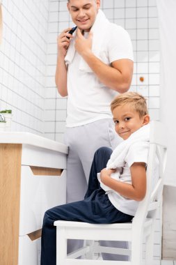 Smiling son looking at camera while sitting on chair near happy father shaving with electric razor in bathroom clipart