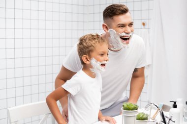 Baba ve oğul, banyo malzemeleriyle lavaboya yaslanıp gülerken, yüzlerinde tıraş köpüğü olan bir baba ve oğul.