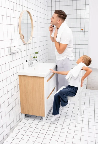儿子坐在靠近父亲的椅子上 用电动剃须刀刮胡子 看着浴室里的镜子 — 图库照片