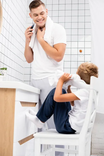 Alegre Padre Sosteniendo Afeitadora Eléctrica Mirando Hijo Sentado Silla Baño — Foto de Stock