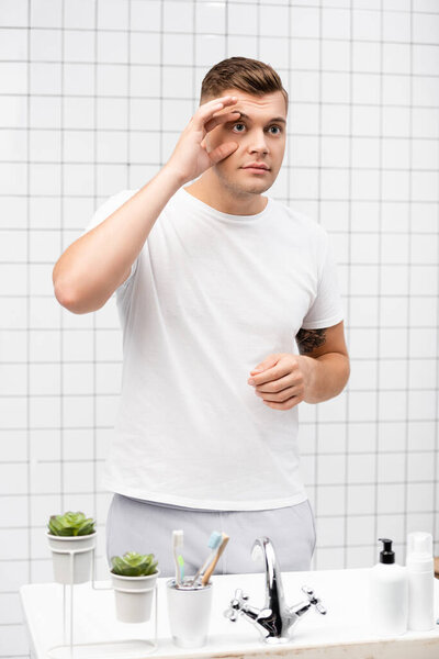 Young adult man touching skin around eye while standing near sink in bathroom