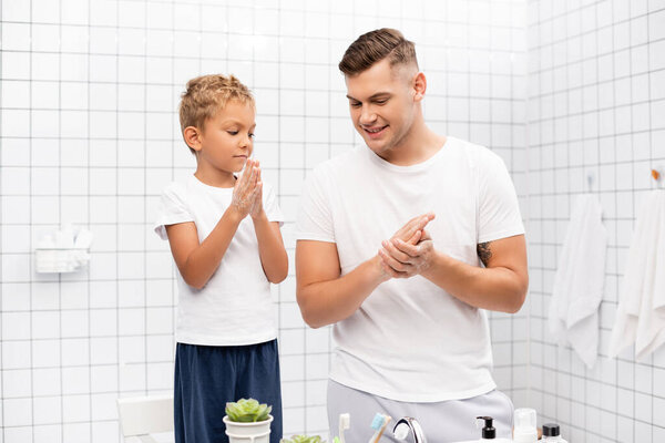 Son rubbing soap between hands, while standing on chair near smiling father in bathroom