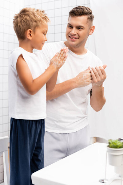 Smiling father looking at son rubbing soap between hands while standing on chair near washbasin in bathroom