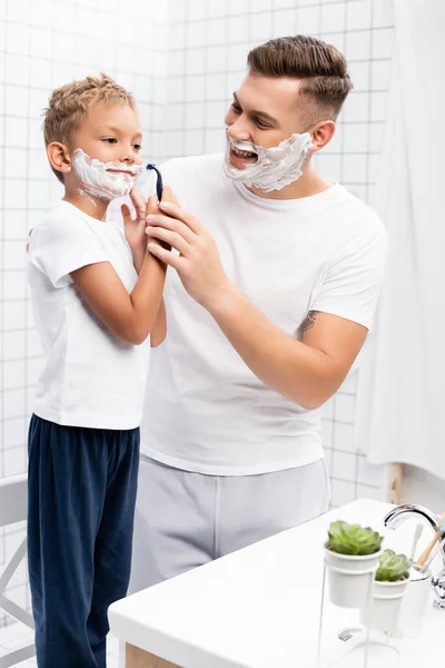 Smiling father looking at son with shaving foam holding safety razor near cheek while standing on chair in bathroom