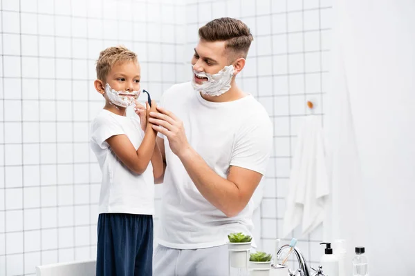 Happy father looking at son with shaving foam on face holding safety razor near cheek while standing on chair in bathroom