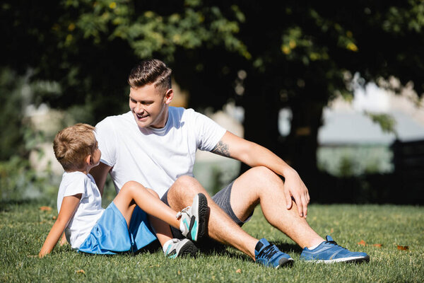 Cheerful father in sportswear looking at son, while sitting on grass in park on blurred background