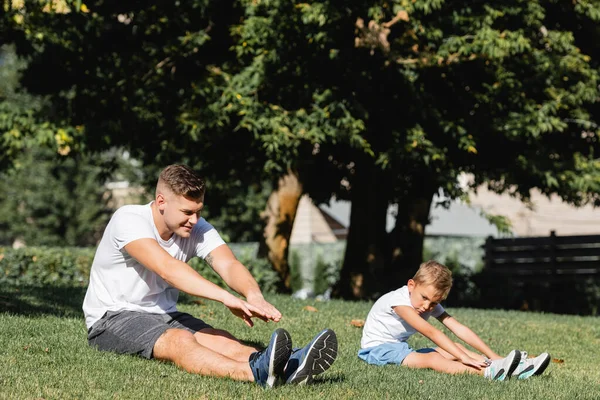 Apa Fia Sportruházatban Kinyújtott Kézzel Bemelegítenek Miközben Füvön Ülnek Homályos — Stock Fotó