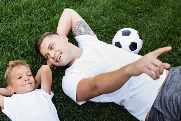 Overhead view of happy father pointing with finger, while looking at smiling son lying with hands behind head on grass