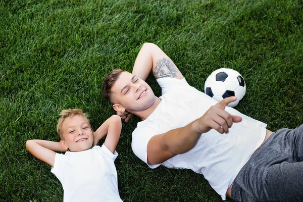 草の上に頭の後ろの手で息子の近くに横たわっている間 指で指して笑みを浮かべて父親の概要 — ストック写真