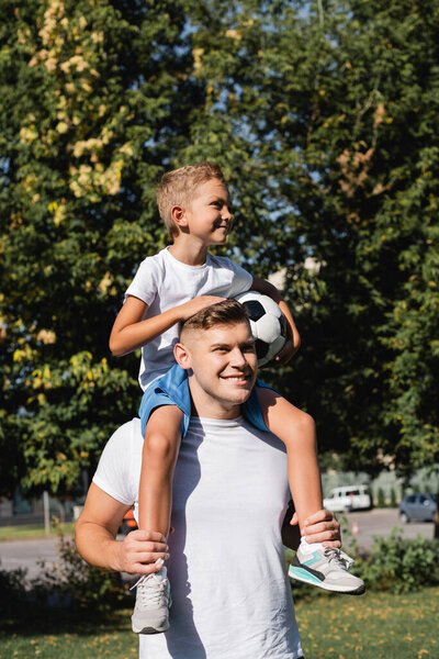 Happy son holding ball and riding piggyback on smiling father in park on blurred background 