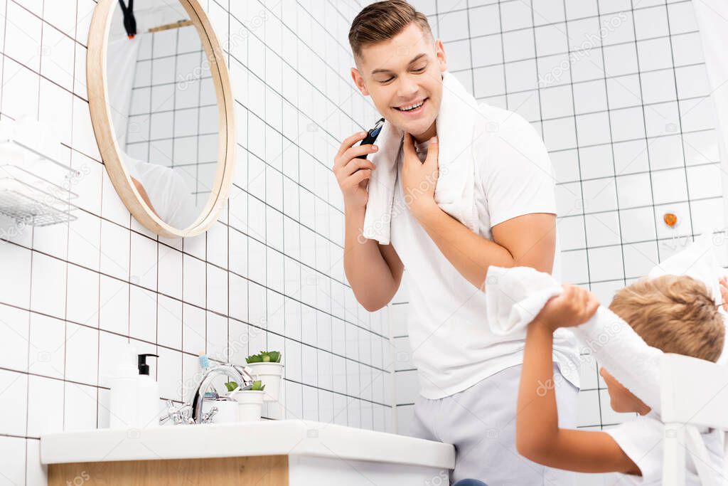 Happy father shaving with electric razor and looking at son sitting on chair in bathroom