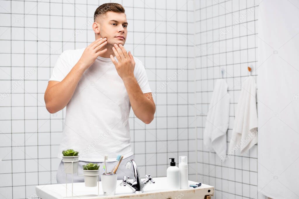 Young adult man touching face while standing near sink with toiletries in bathroom