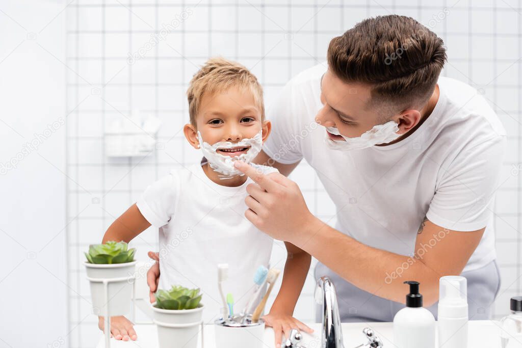 Smiling father hugging and touching chin of son with shaving foam on face in bathroom