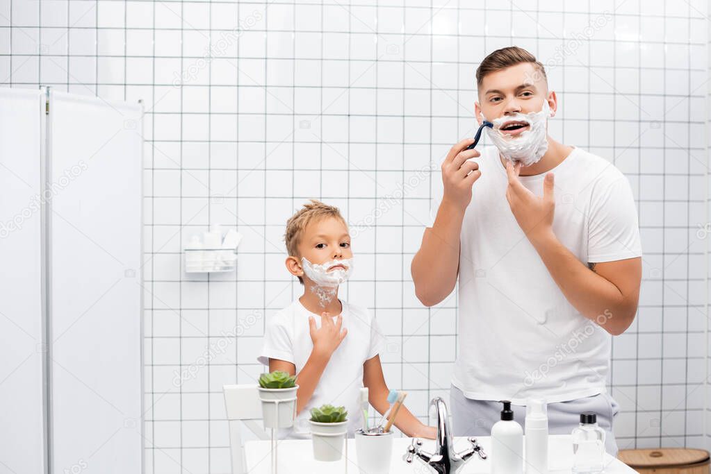 Focused man using safety razor while standing near boy with shaving foam on face in bathroom