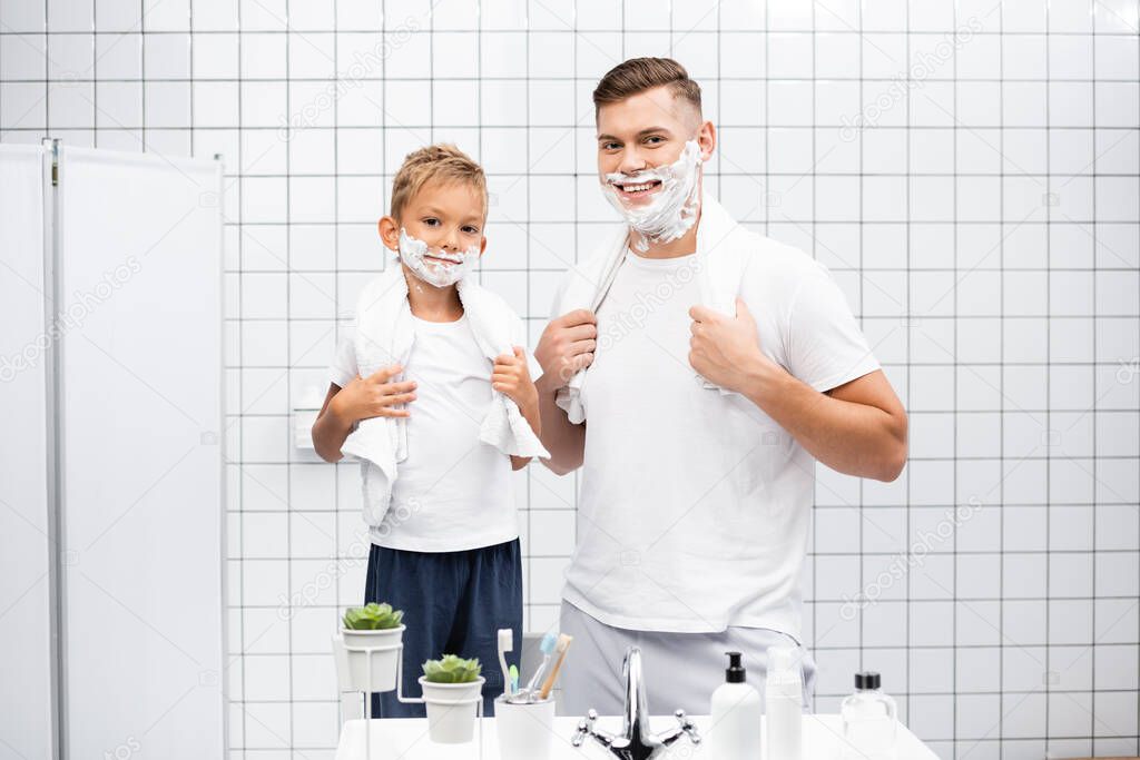 Happy son and father with shaving foam on faces, touching towels over necks while standing in bathroom