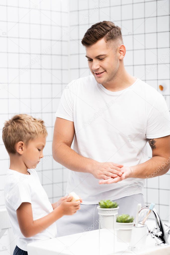 Positive father washing hands and looking at son with soap standing near washbasin in bathroom