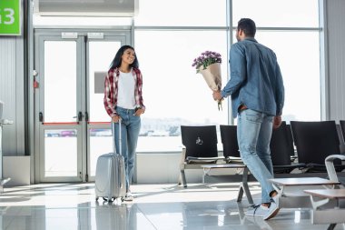 man holding wrapped flowers while meeting cheerful african american girlfriend in airport clipart