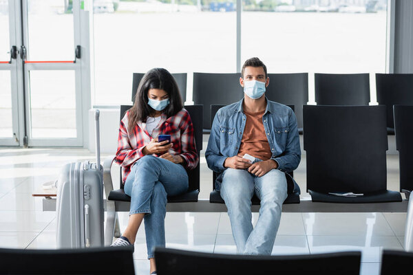 interracial couple in medical masks sitting and using phones near luggage in departure lounge 