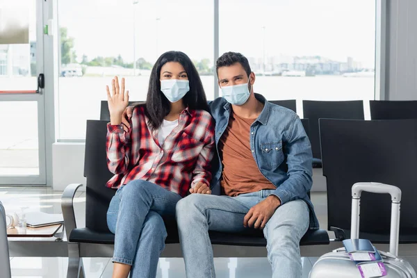 african american woman waving hand near man in medical mask near luggage in departure lounge