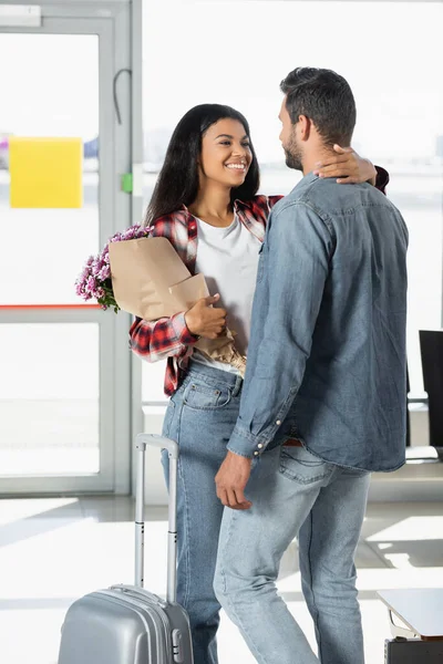 Feliz Afro Americana Segurando Flores Embrulhadas Abraçando Namorado Aeroporto — Fotografia de Stock