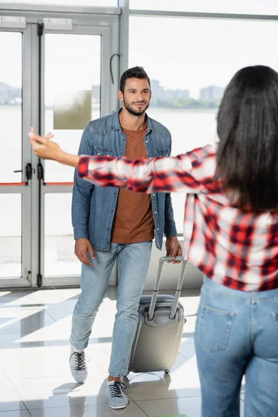 cheerful man walking with luggage while looking at african american woman with outstretched hands on blurred foreground