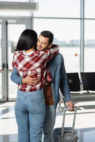 cheerful man smiling while hugging african american woman in airport 