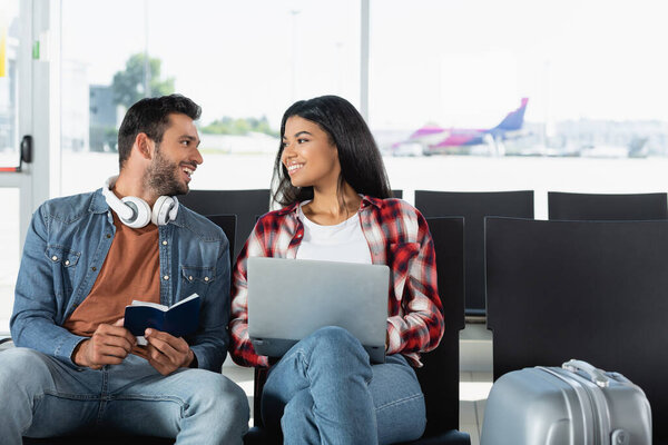 happy interracial couple looking at each other near laptop in airport 