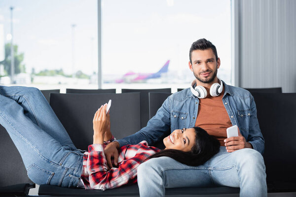 счастливая африканская американская женщина с помощью телефона и лежа на бородатый мужчина в зале вылета 