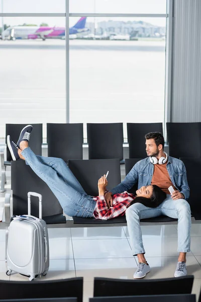 happy african american woman holding phone and lying on bearded man sitting near baggage in departure lounge