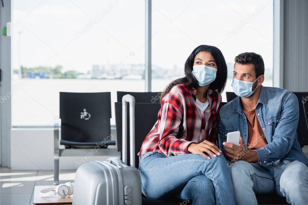 interracial couple in medical masks sitting near luggage in departure lounge 
